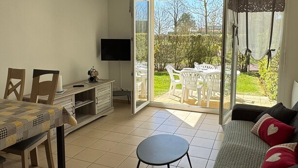 A room with a dining table, chairs, a sofa, a TV, and a sliding glass door leading to an outdoor patio with a table and chairs.