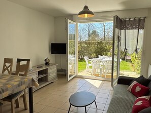 Et værelse med et spisebord, stole, en sofa, et fjernsyn og en skydedør til en udendørs terrasse med et bord og stole.