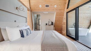 A modern bedroom with a large bed, a wooden ceiling, and a sliding glass door leading to a bathroom with a bathtub. - Cedar Treehouse @ A-Frame Ranch (Fredericksburg)