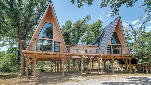A wooden cabin with a triangular roof, large windows, and a wooden deck. - Cedar Treehouse @ A-Frame Ranch (Fredericksburg)