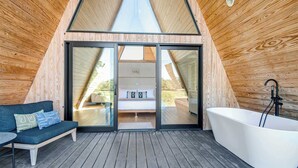 A modern, wooden-paneled room with a large sliding glass door leading to a bedroom, a white bathtub, and a dark blue sofa with cushions. - Cedar Treehouse @ A-Frame Ranch (Fredericksburg)