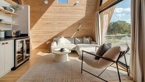 A modern living room with a sofa, coffee table, and chair, wooden ceiling, and large windows. - Cedar Treehouse @ A-Frame Ranch (Fredericksburg)
