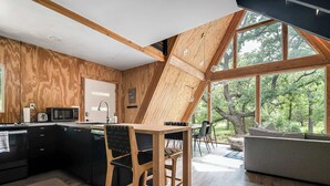 A modern kitchen with dark cabinetry, a wooden dining table, and a large window overlooking a forest. - Large A-Frame in Nature - minutes from Main (Fredericksburg)