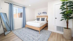 A bedroom with a bed, a large window with curtains, a bedside table, a potted plant, and artwork on the wall. - 4 Miles to Beach Central and Newly Renovated (San Diego)