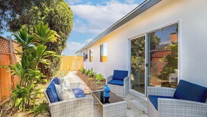 A patio area with wicker furniture, a glass table, and a view of a house with a wooden fence and plants. - 4 Miles to Beach Central and Newly Renovated (San Diego)