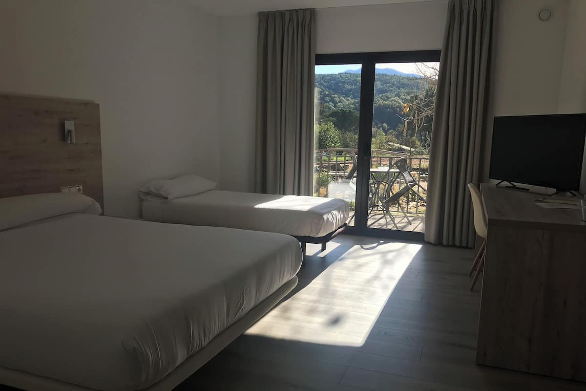 Habitación de hotel con dos camas, un escritorio y vistas a un balcón con mobiliario exterior.