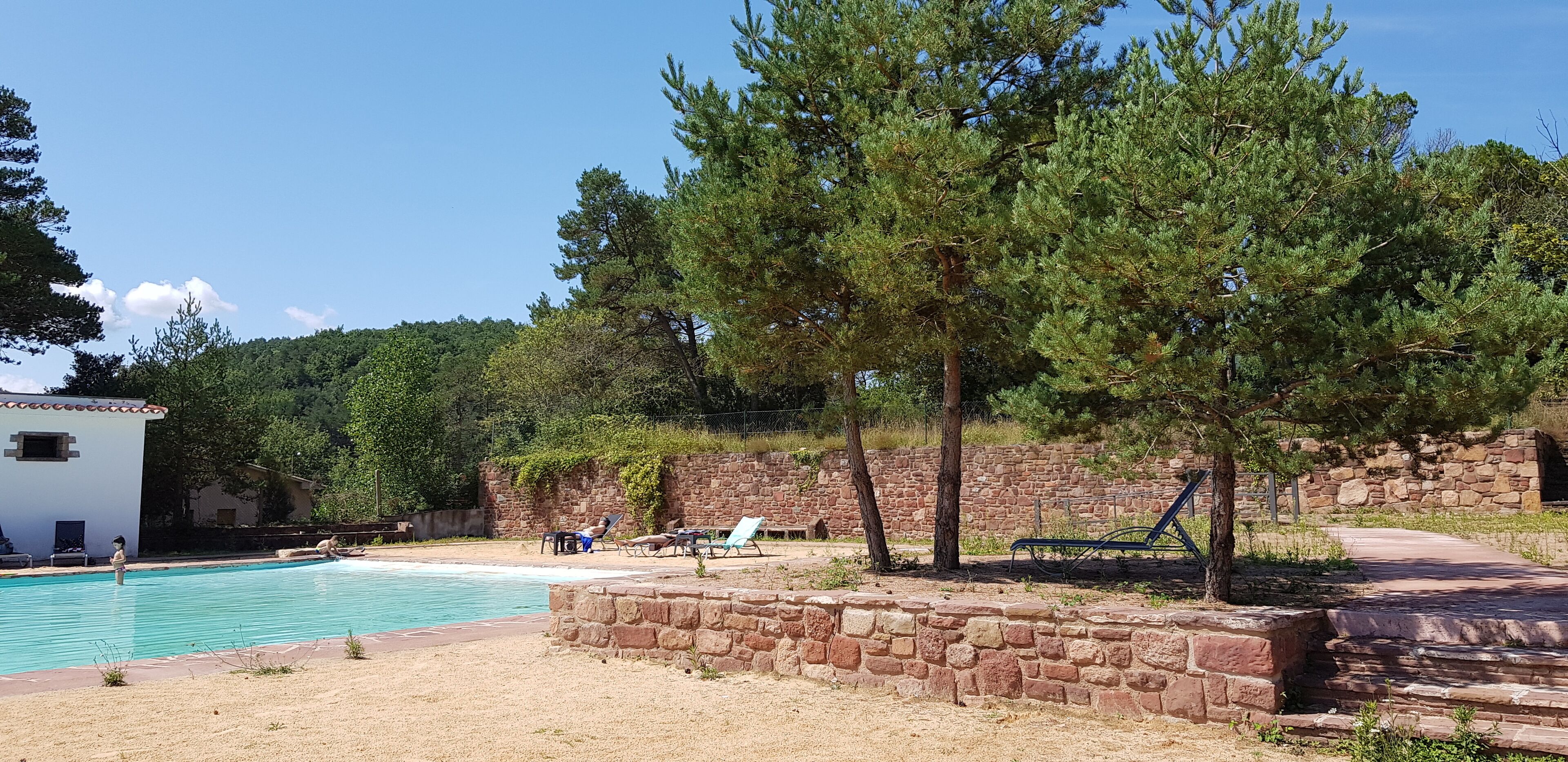 Zona de piscina con sillas de descanso, un muro de piedra y un edificio de fachada blanca.