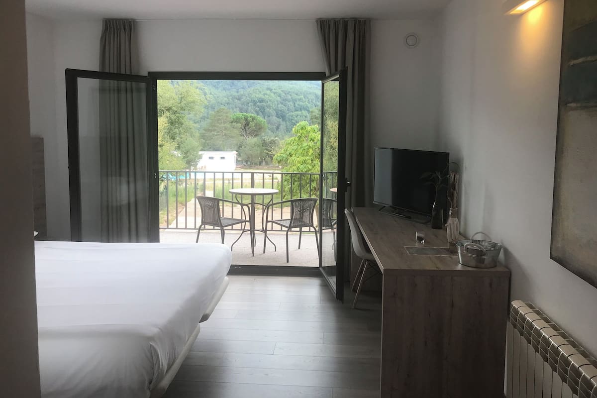 Habitación de hotel con cama, escritorio con televisor, pequeña mesa y sillas. La habitación tiene balcón con vista a zonas verdes y un edificio.
