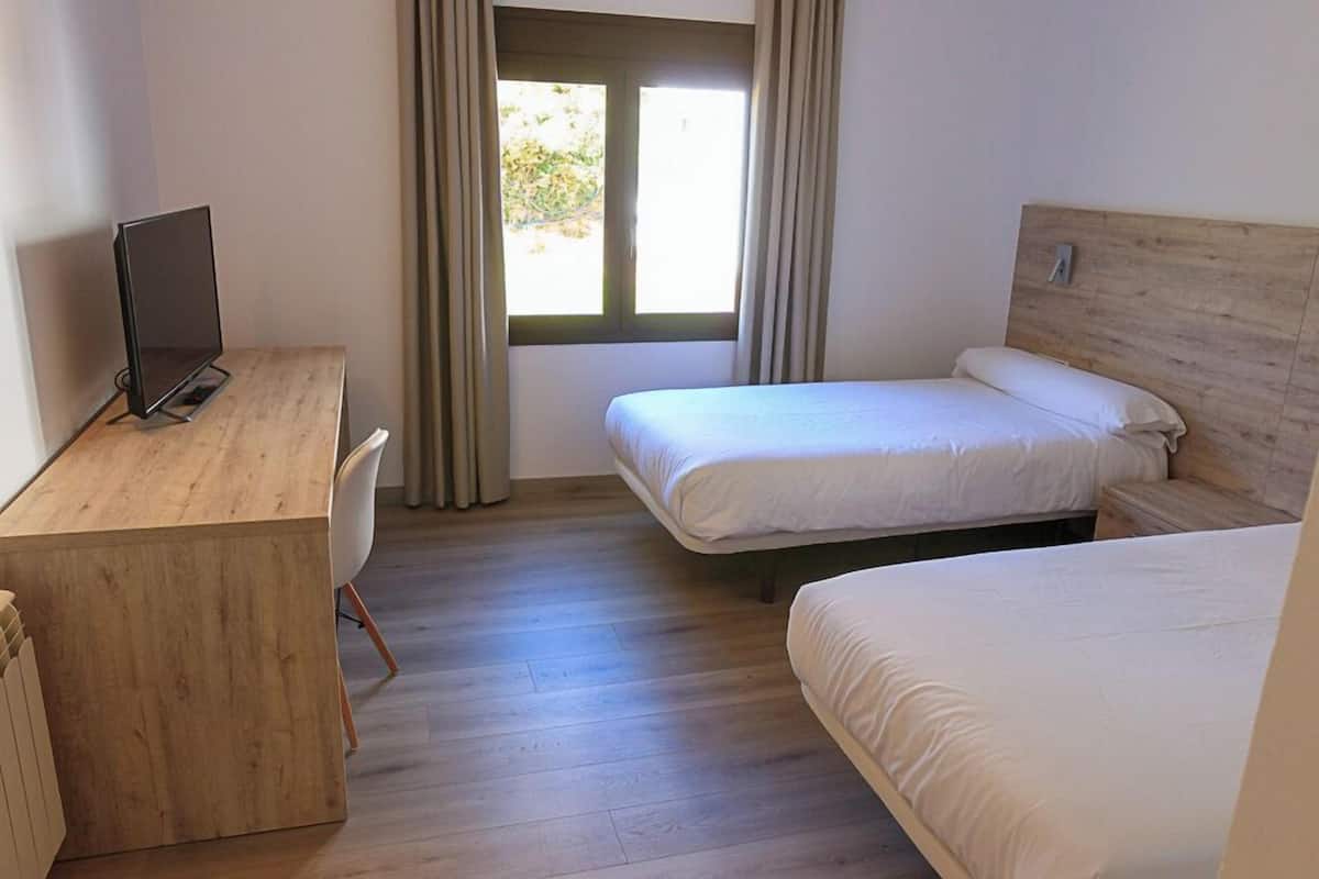 Una habitación con un escritorio de madera, dos camas individuales, una ventana con cortinas y vistas a zonas verdes.