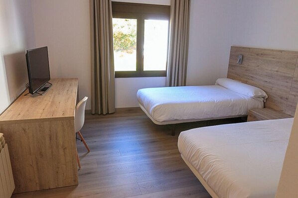 A room with a wooden desk, two single beds, a window with curtains, and a view of greenery outside. - HOSTAL LA GUINEU (Viladrau)