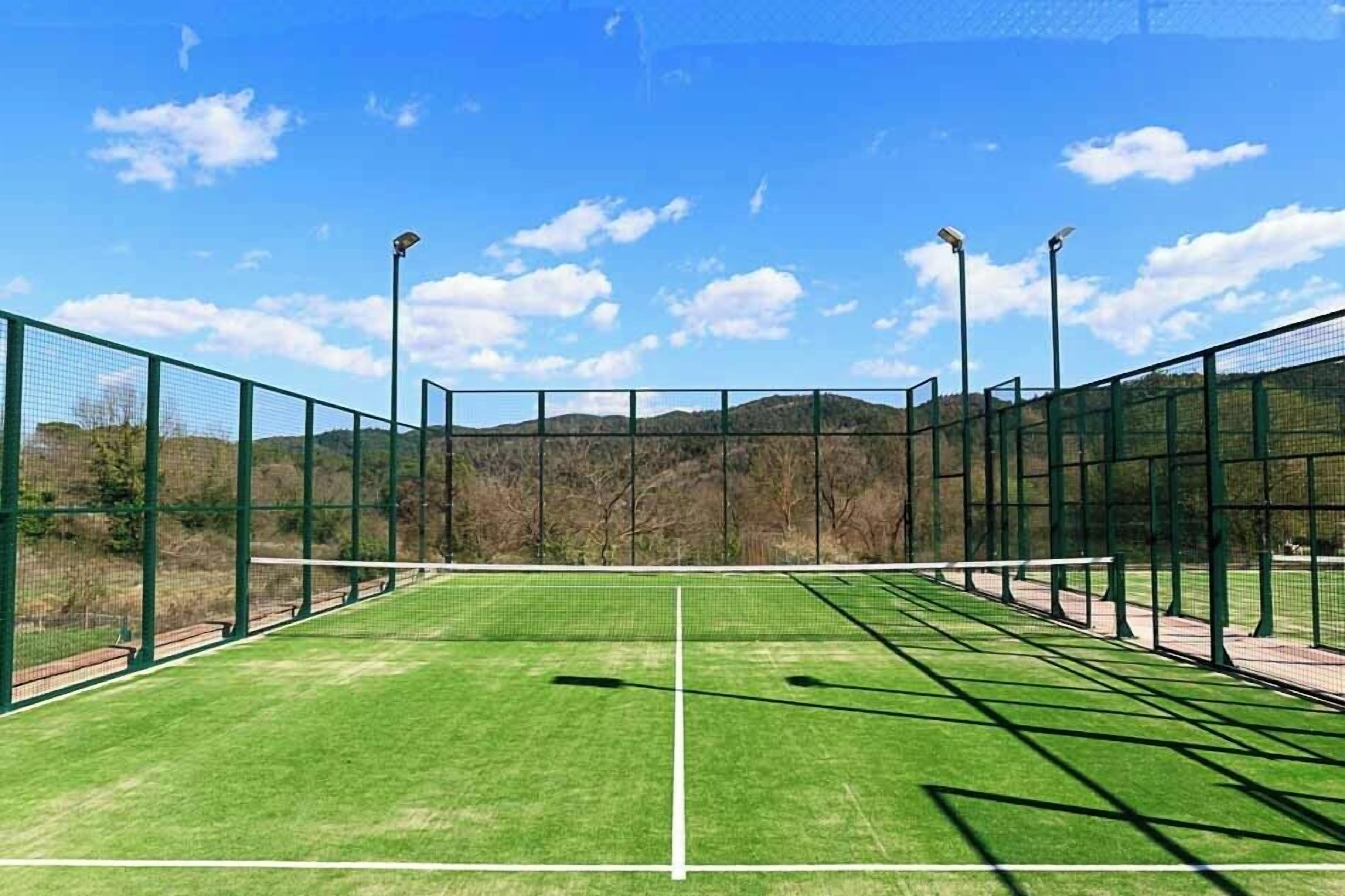 A tennis court with green artificial turf, surrounded by a green fence and equipped with floodlights.