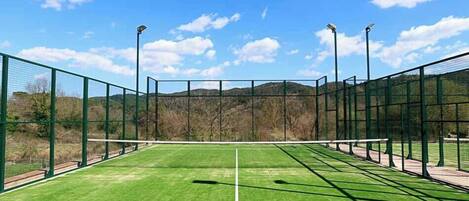 A tennis court with green artificial turf, surrounded by a green fence and equipped with floodlights.