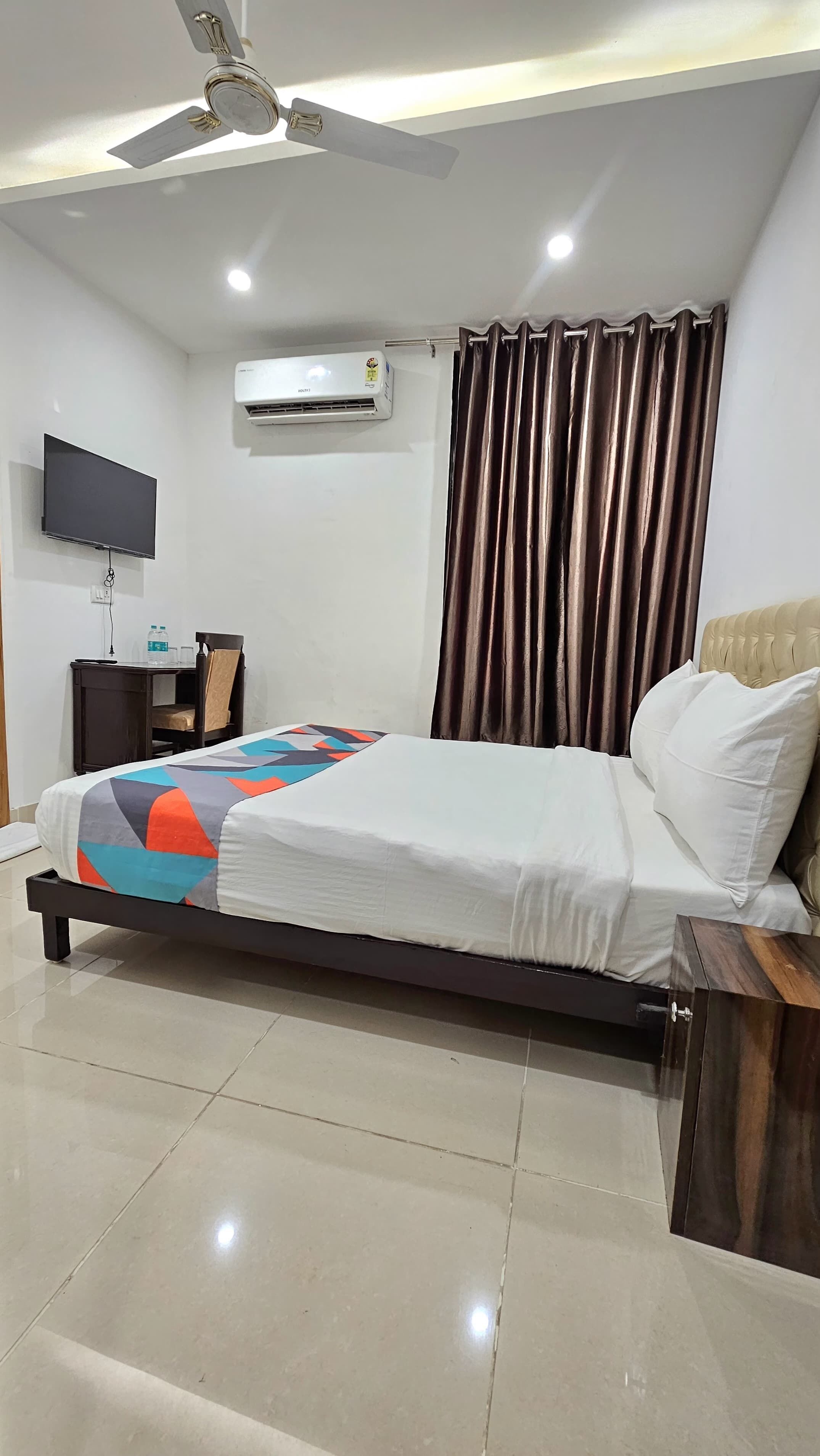 A bedroom with a bed, a flat-screen TV, an air conditioning unit, a ceiling fan, a desk with a chair, and a window with curtains.