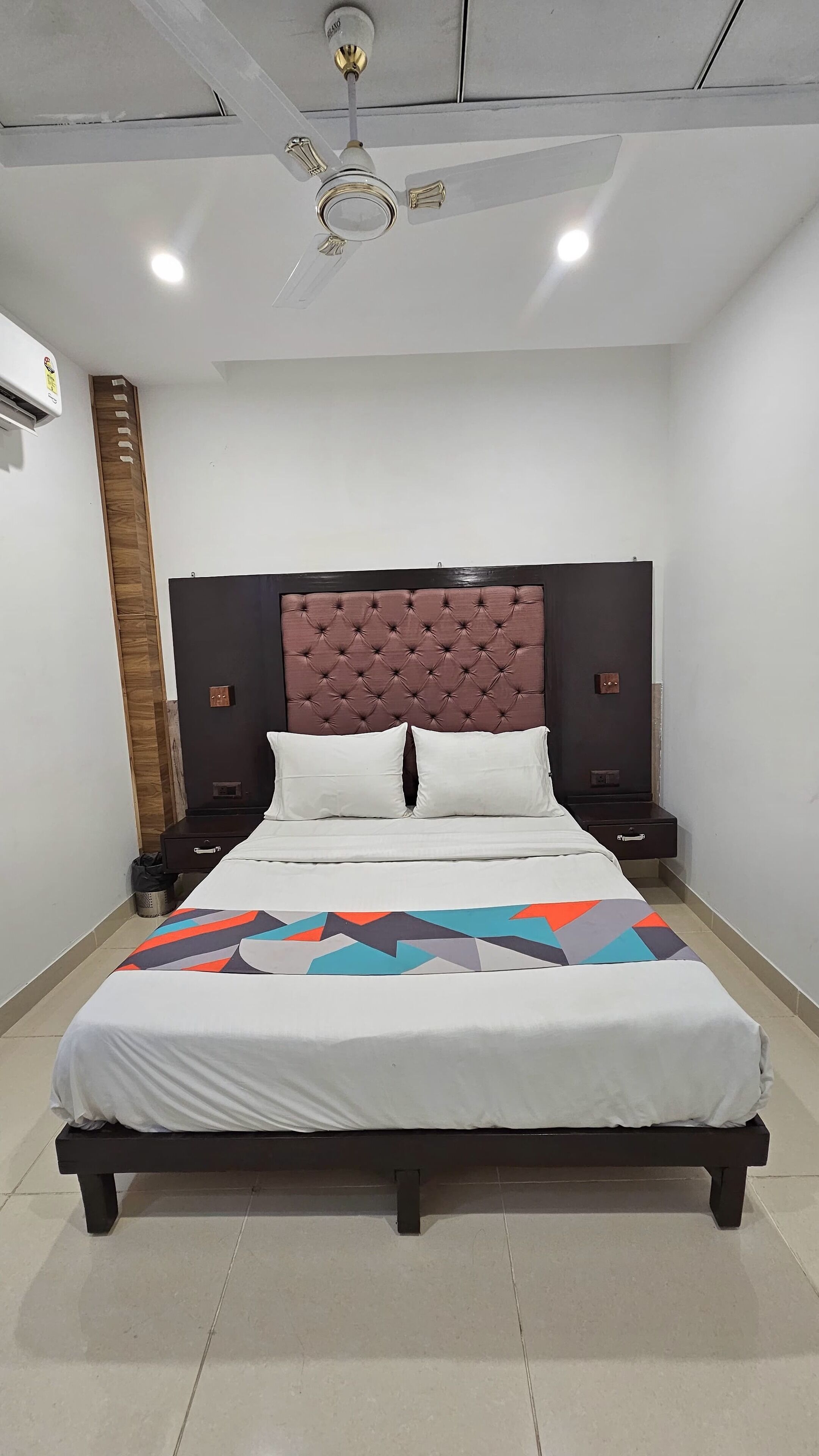A modern bedroom with a tufted headboard, a bed with a colorful geometric-patterned bedspread, two bedside tables, and a ceiling fan.