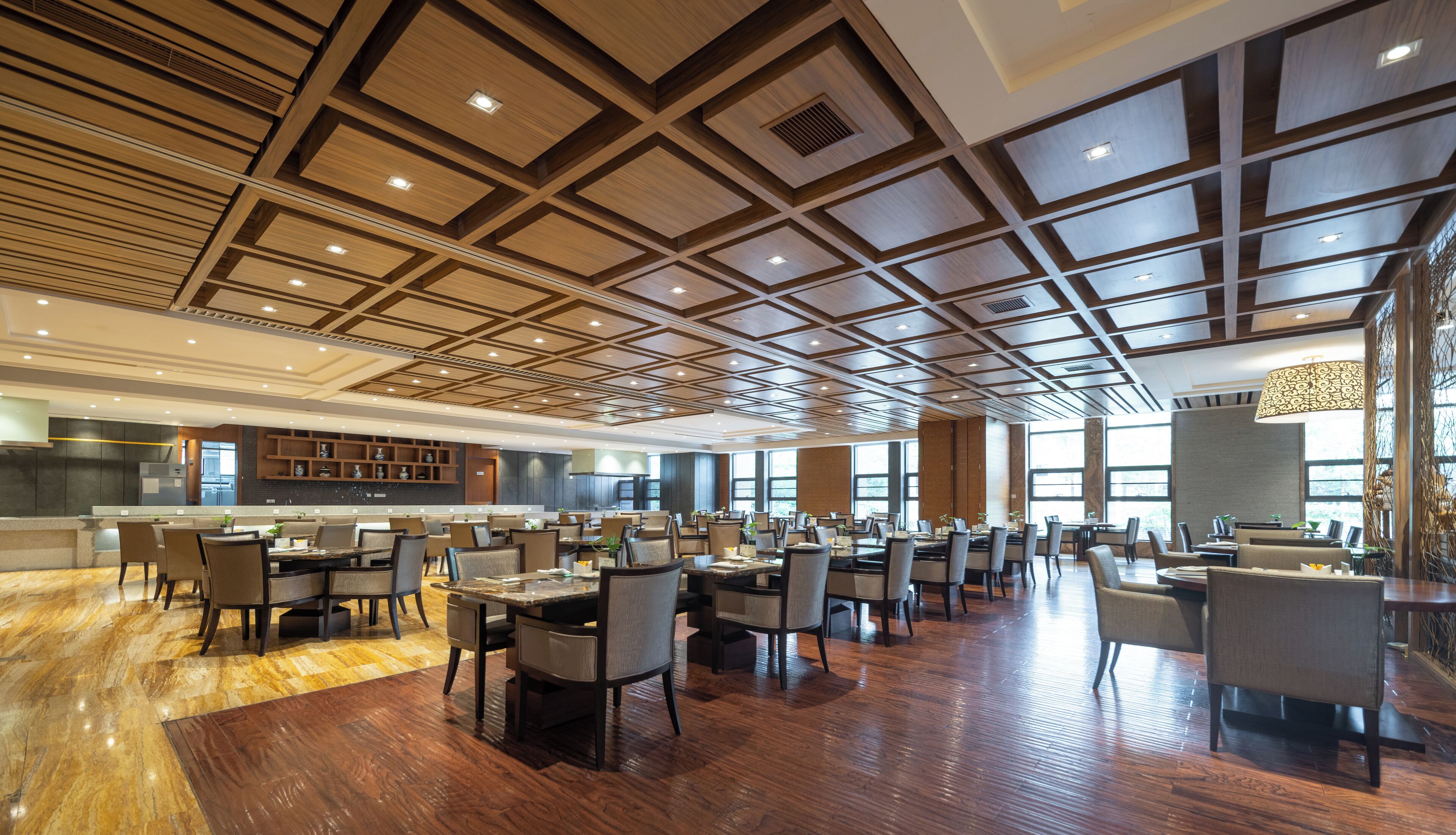 A spacious dining area with multiple tables and chairs, a modern kitchen in the background, and a high ceiling with geometric patterns.