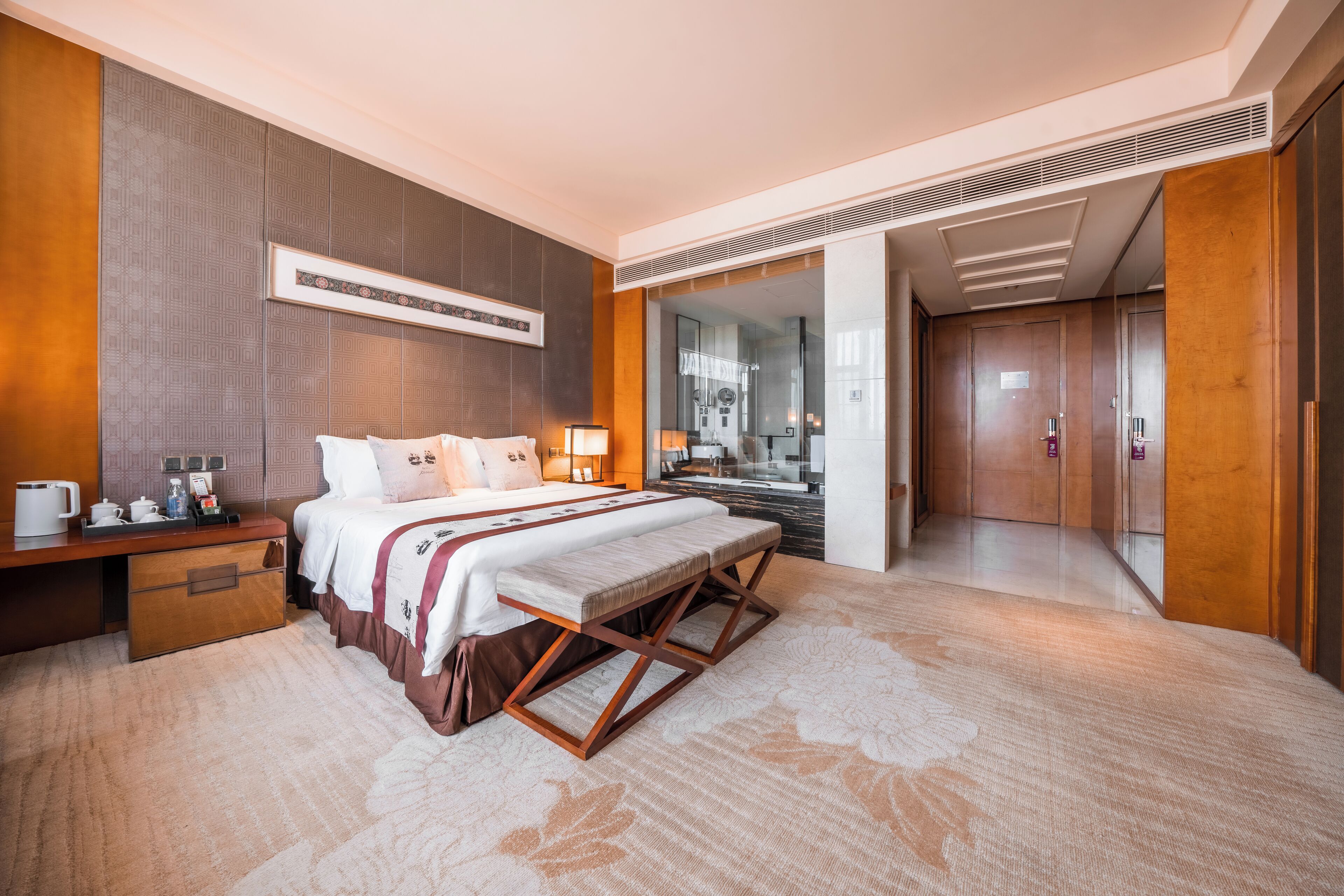 Habitació d'hotel moderna amb un llit gran, taules de nit, escriptori i un bany visible a través d'una porta de vidre.