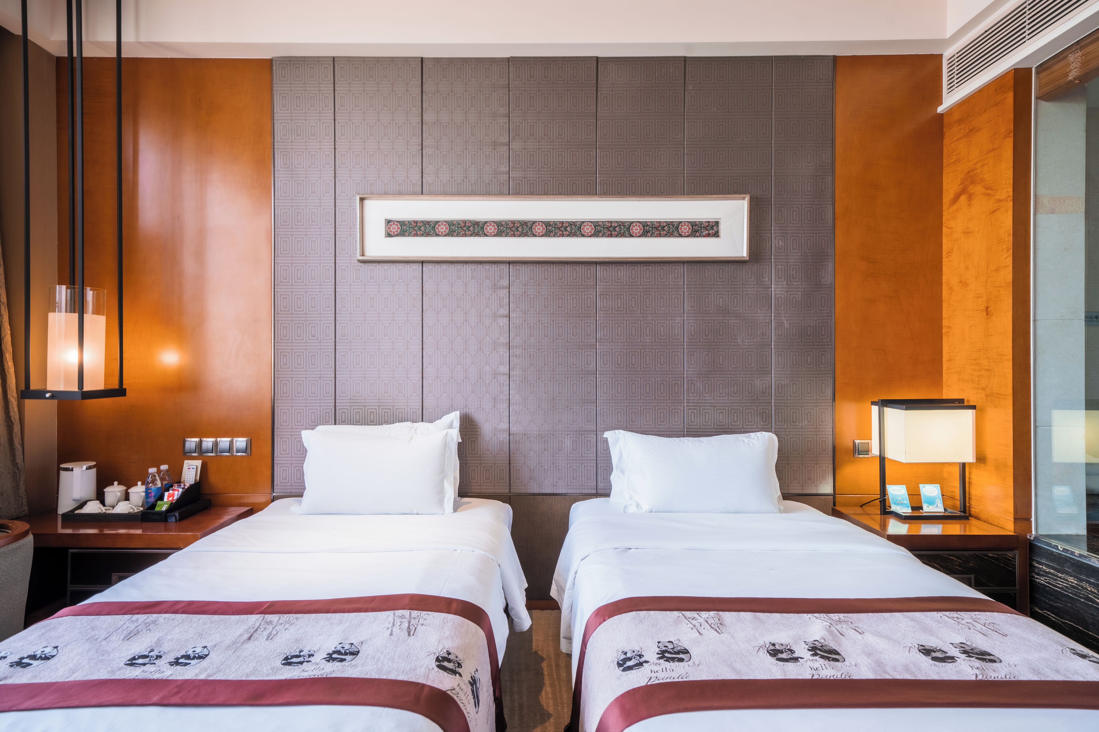 A hotel room with two beds, a framed artwork above the beds, a nightstand with a lamp, and a desk with a phone and toiletries.