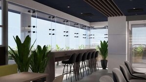Un espacio interior moderno con una mesa larga y varias sillas, ventanas amplias y plantas en macetas.