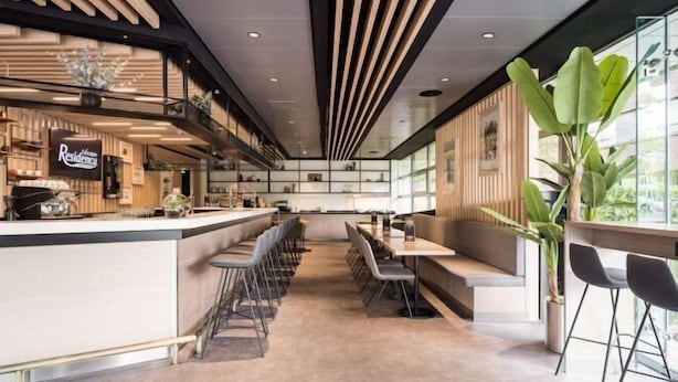 Un restaurante moderno con una barra alargada, asientos tipo bar y un techo alto con iluminación lineal.