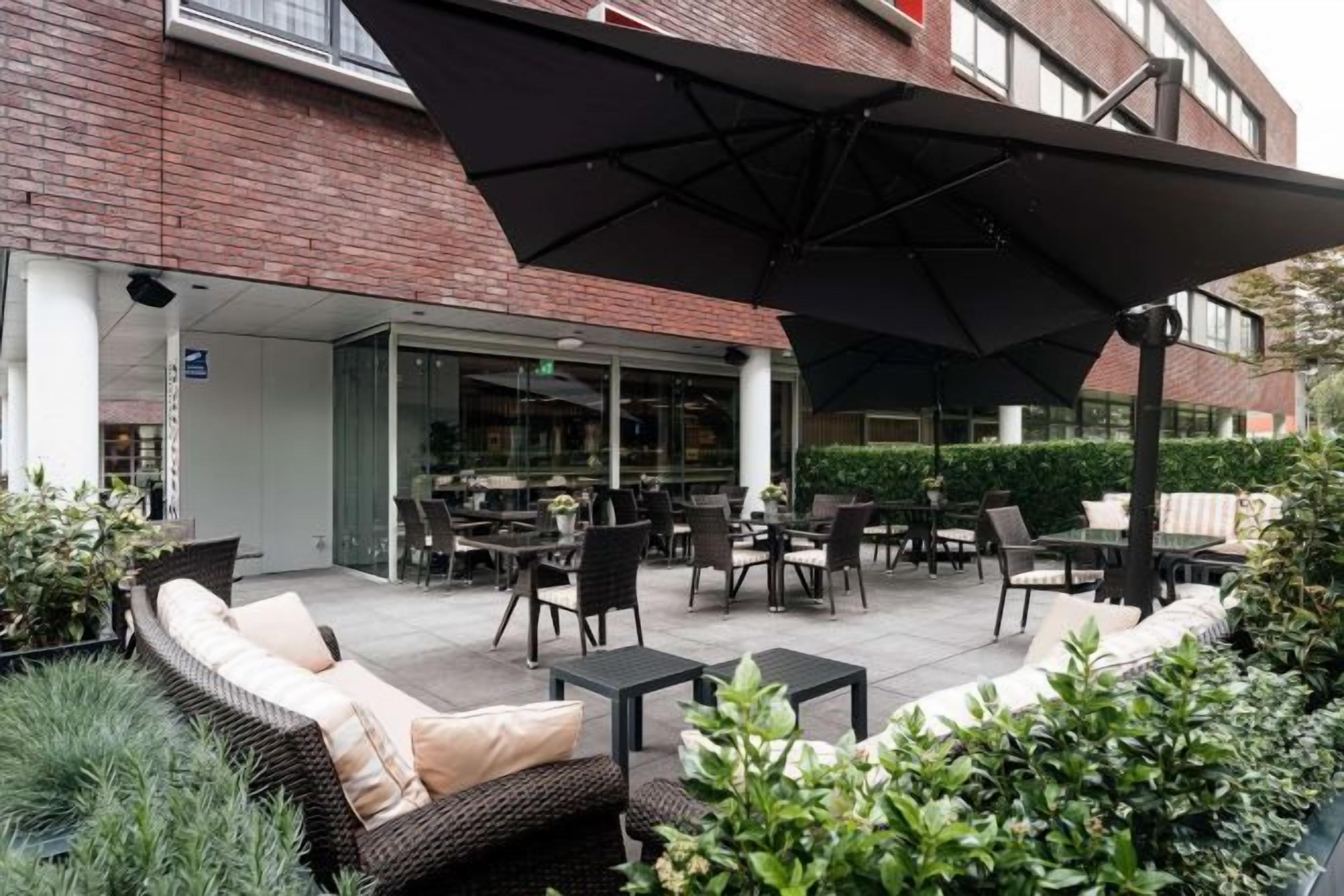 Un patio al aire libre con mobiliario de mimbre, un paraguas grande y un edificio de ladrillo al fondo.