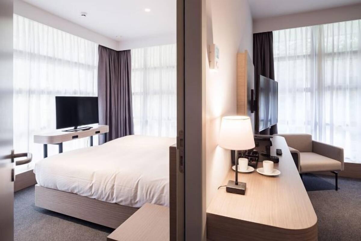 Quarto de hotel com uma cama, uma escrivaninha com candeeiro, uma televisão e uma cadeira.