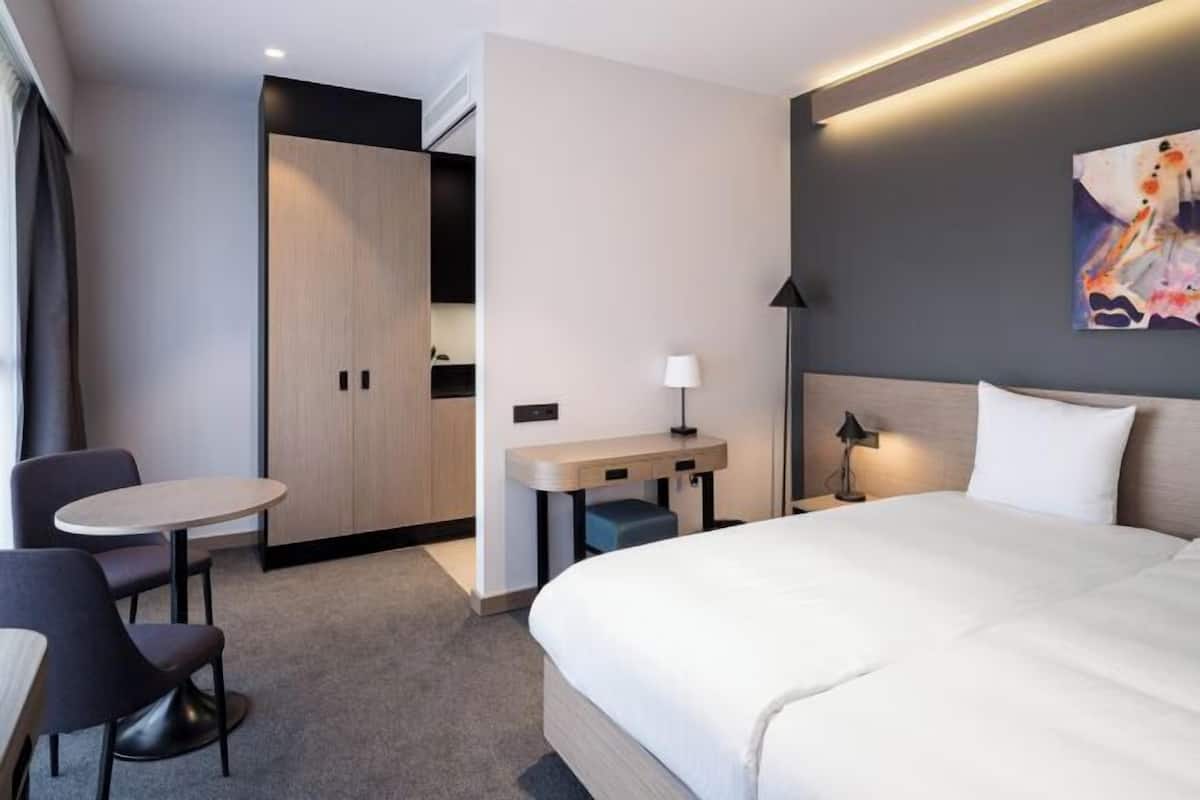 Quarto de hotel com cama, escrivaninha, cadeira e mesa pequena.