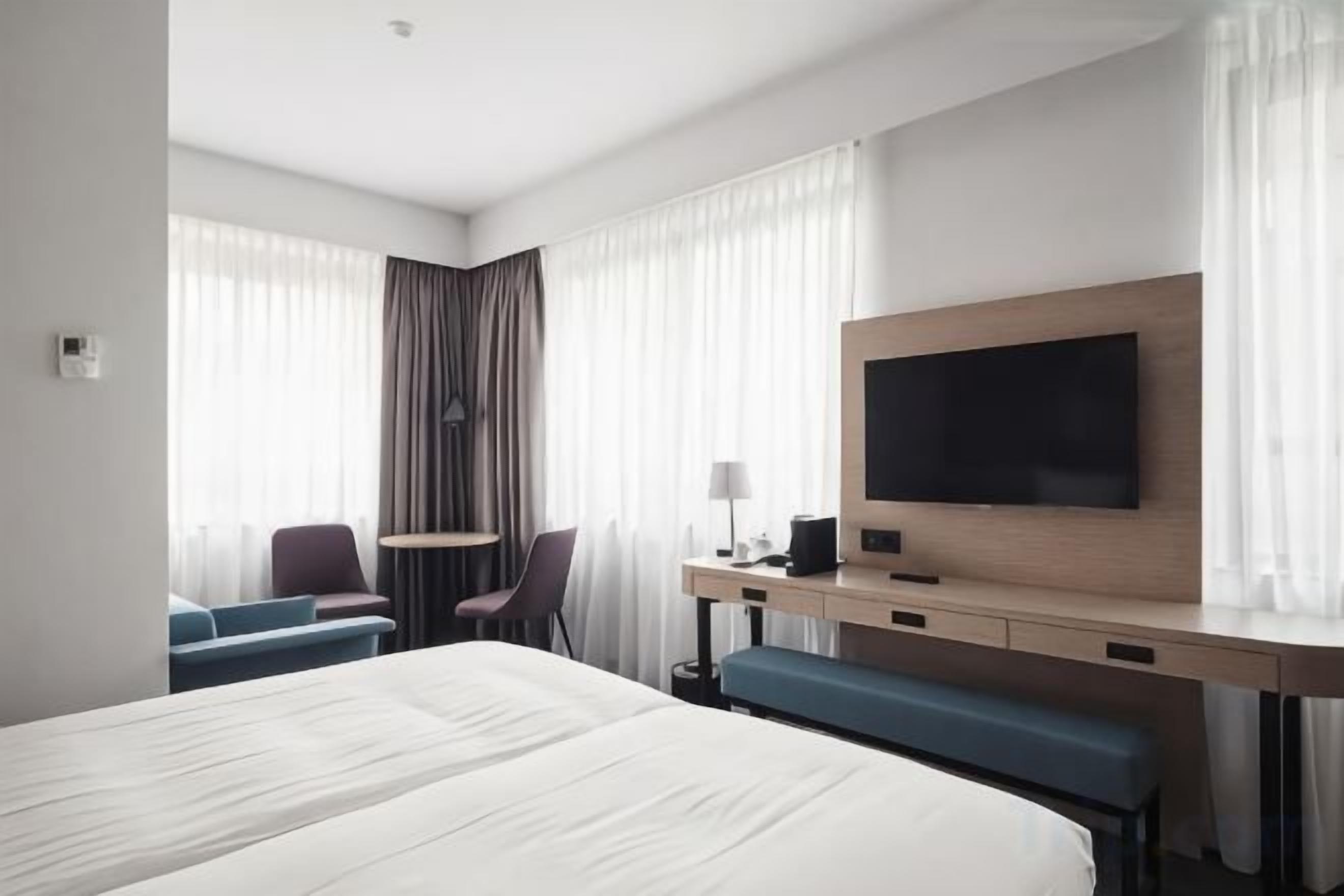 Une chambre d’hôtel avec un grand lit, un bureau doté d’un téléviseur à écran plat, une chaise, une petite table et un banc.