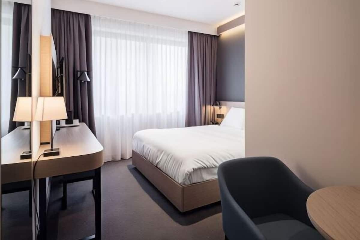 Quarto de hotel com uma cama, uma escrivaninha com candeeiro, uma cadeira e uma pequena mesa.