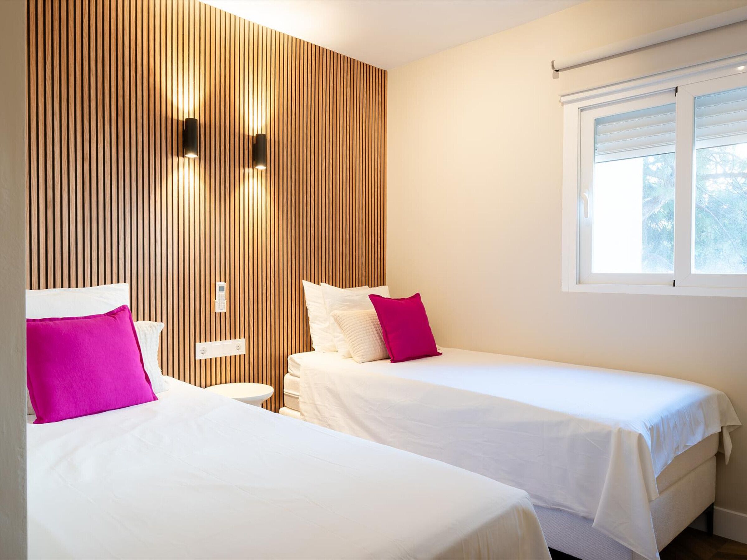 A hotel room with two beds, wooden paneling on one wall, a window with blinds, and a small bedside table with a lamp.