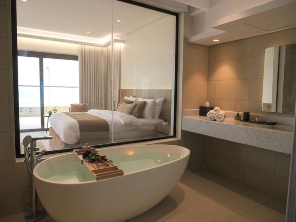 A modern bathroom with a large bathtub, a glass shower enclosure, and a visible bedroom with a bed and curtains.
