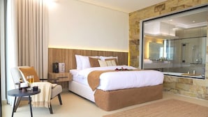A modern hotel room with a large bed, a sitting area with a chair and table, a stone accent wall, and a large mirror.