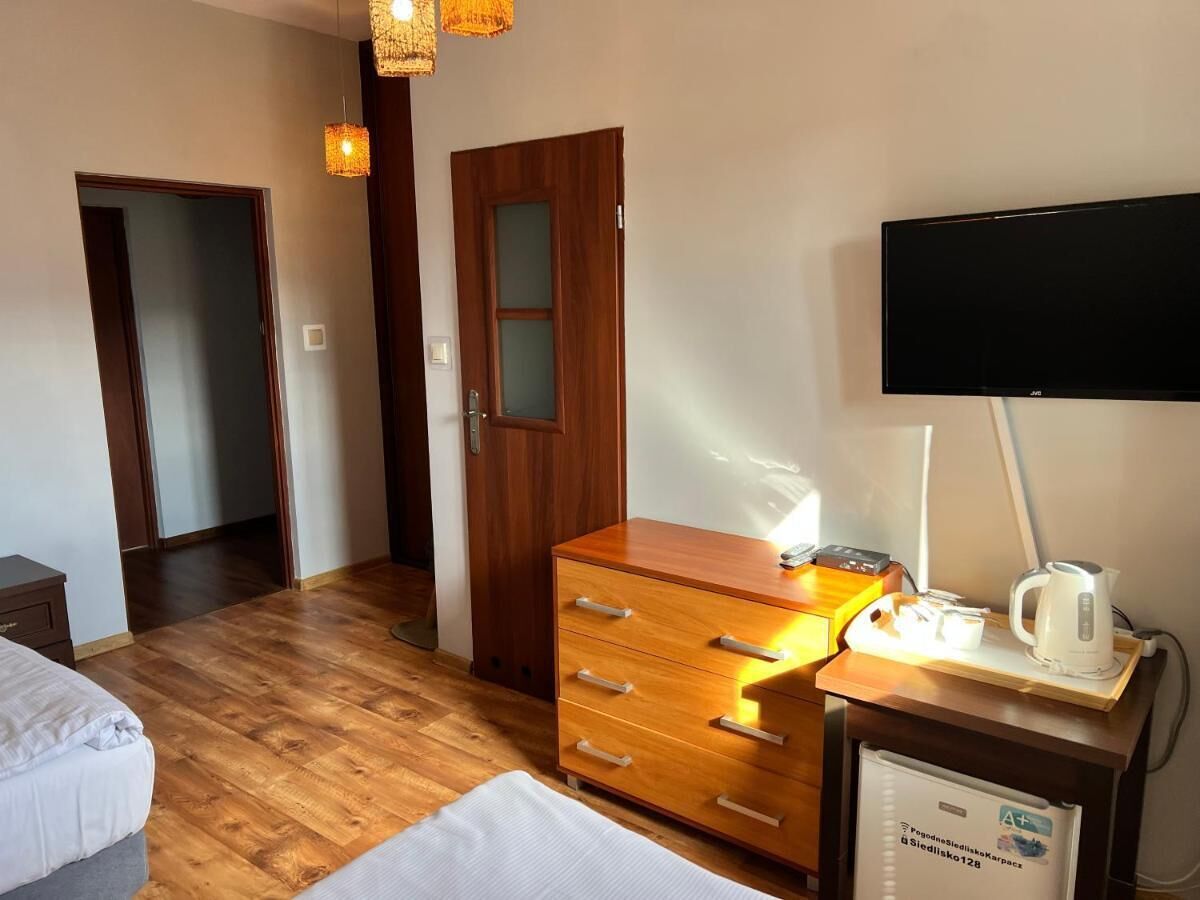 A hotel room with a wooden floor, a wooden dresser with drawers, a flat-screen TV mounted on the wall, a small table with a kettle and cups, and a bed with a white comforter.