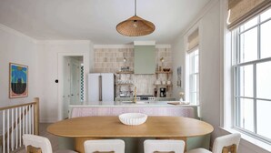 Un coin repas avec une table et des chaises en bois, un luminaire suspendu et, en arrière-plan, une vue sur la cuisine.
