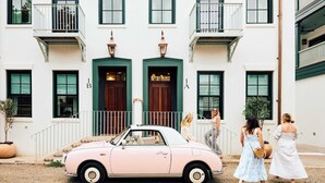 Une voiture ancienne rose est garée devant un bâtiment blanc doté de portes et de balcons verts.
