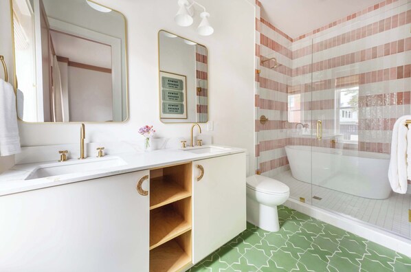 Un baño con lavamanos blanco, detalles dorados, un espejo grande, cabina de ducha con paredes de vidrio y pared con azulejos a rayas.