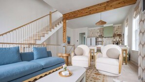 Un salon moderne avec un canapé bleu, des fauteuils blancs, un escalier en bois et une cuisine en arrière-plan.