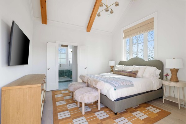 Une chambre à coucher comprenant un lit, une télévision, un bureau en bois, un banc, une table de chevet et une fenêtre donnant sur un paysage agréable.