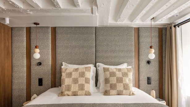 A neatly made bed with checkered pillows, a wooden headboard, and a textured grey wall.