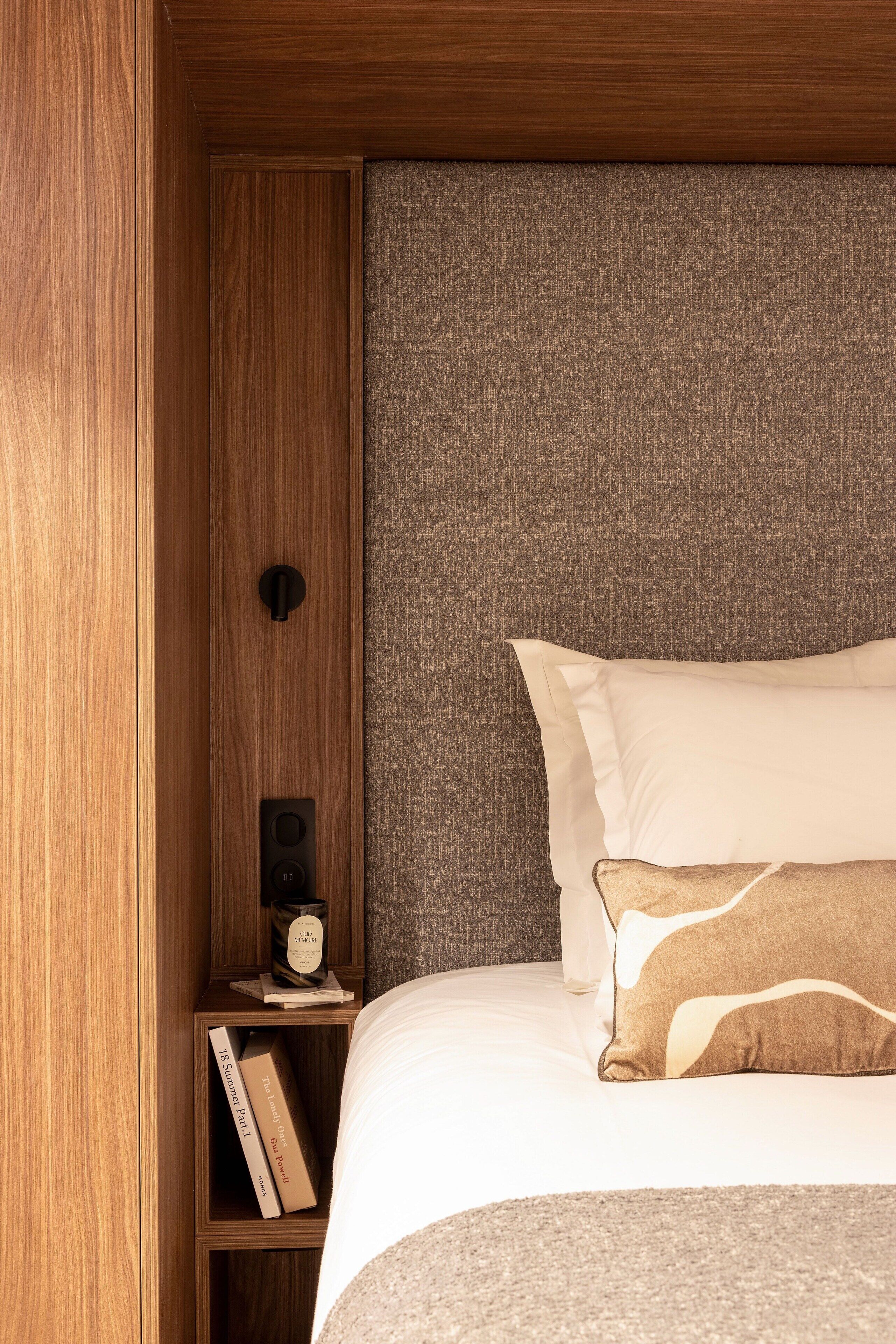 A neatly made bed with white and beige pillows, a wooden nightstand with a book and a small object, and a textured grey headboard.