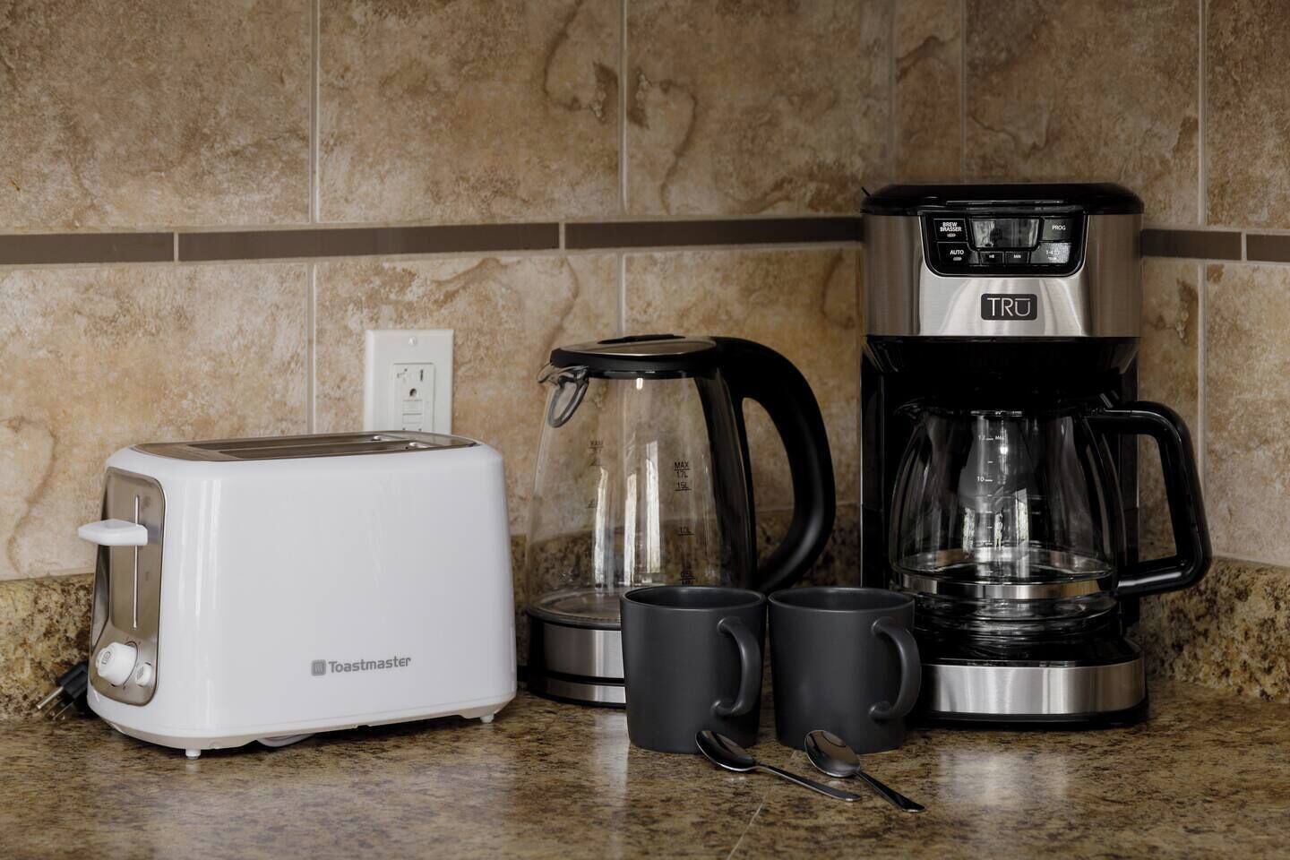 Una cocina con tostadora, cafetera y dos tazas sobre una encimera de granito.
