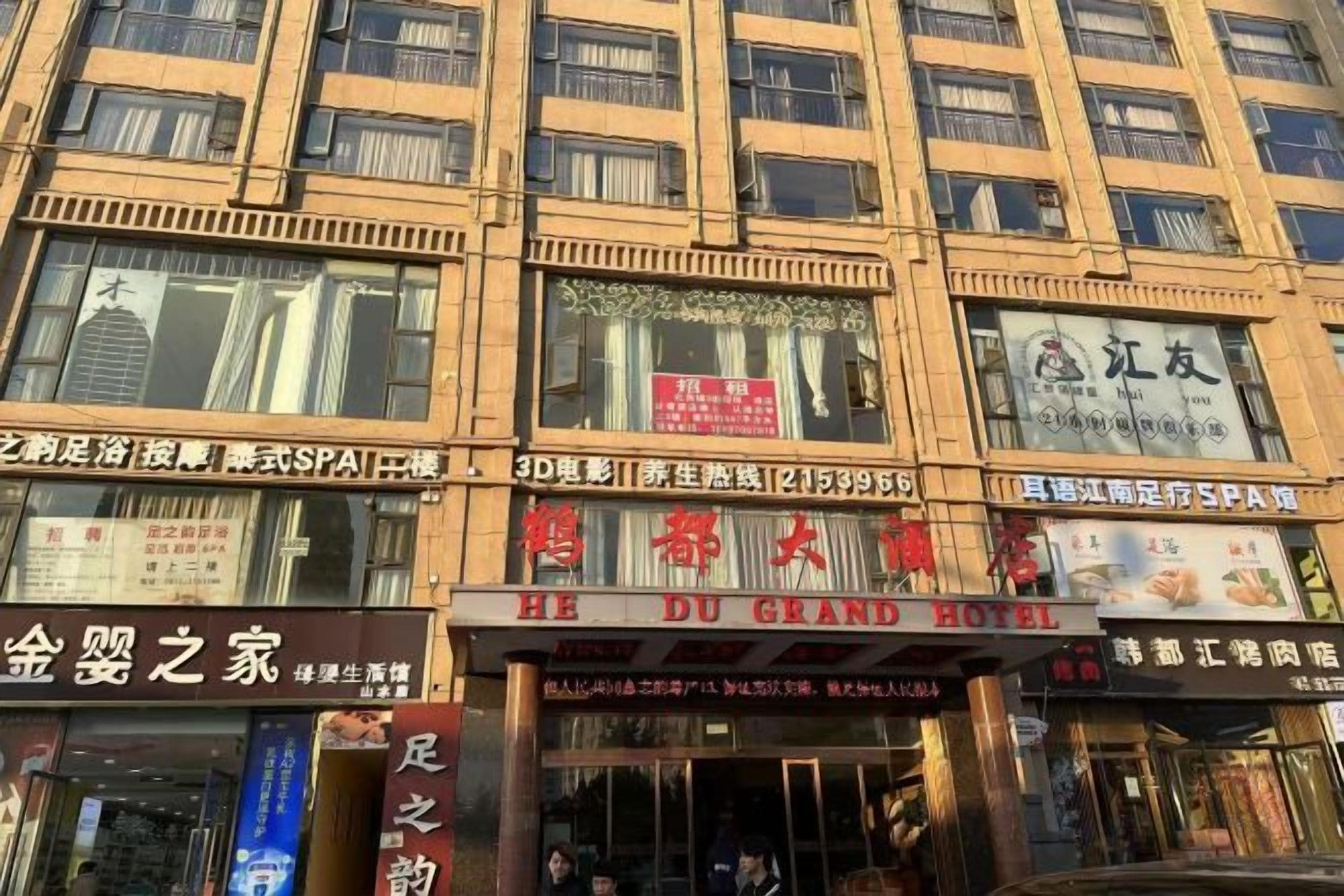En bygning med flere etager, hvor der er forskellige butikker og virksomheder i stueetagen, heriblandt hotellet 'HE DU GRAND HOTEL'.