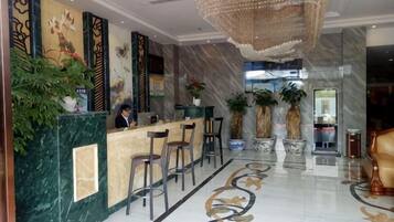 A hotel lobby with a reception desk, bar stools, potted plants, and a chandelier.