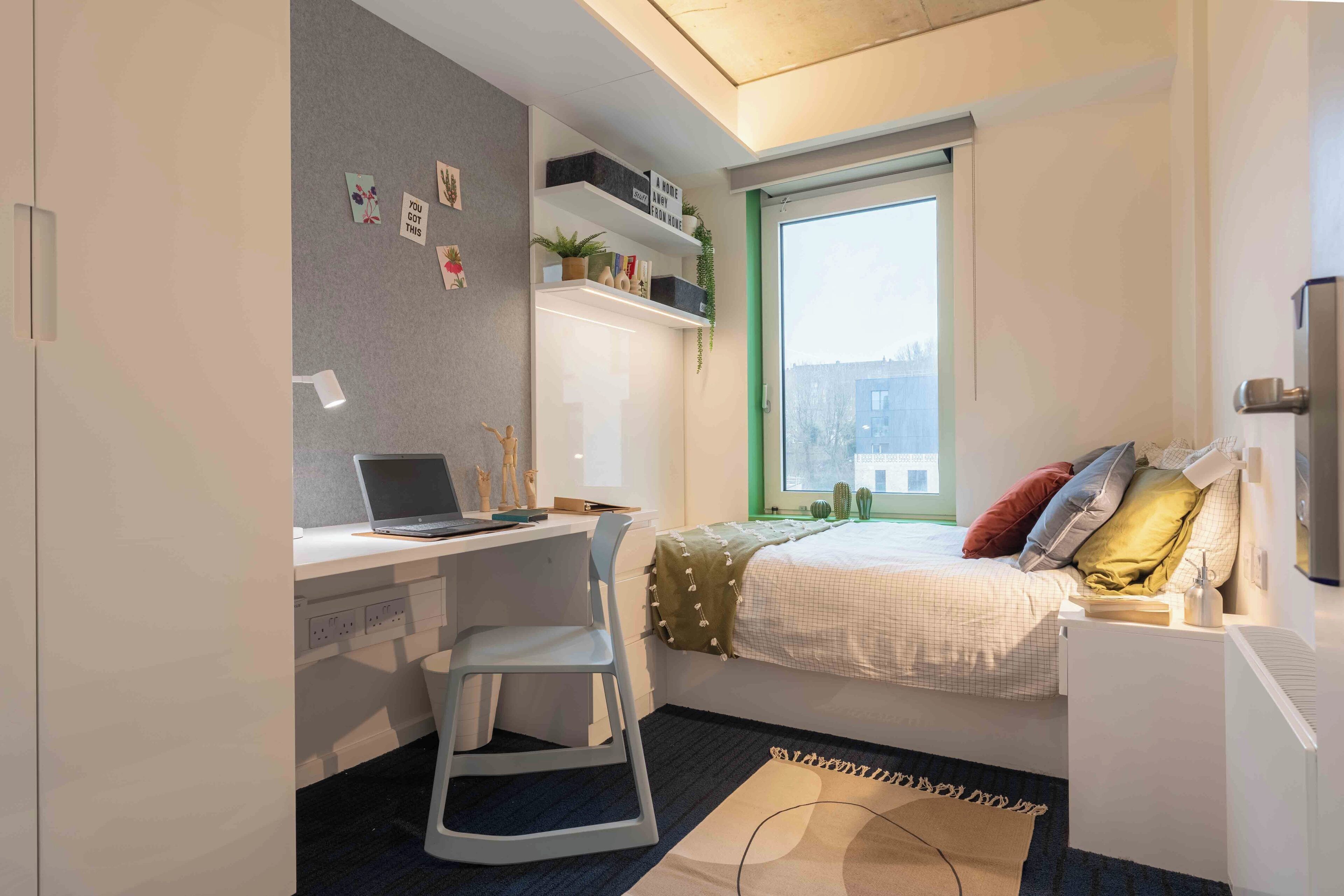 A compact bedroom with a bed, a desk, and a chair. There is a window providing natural light, and a shelf with decorative items.