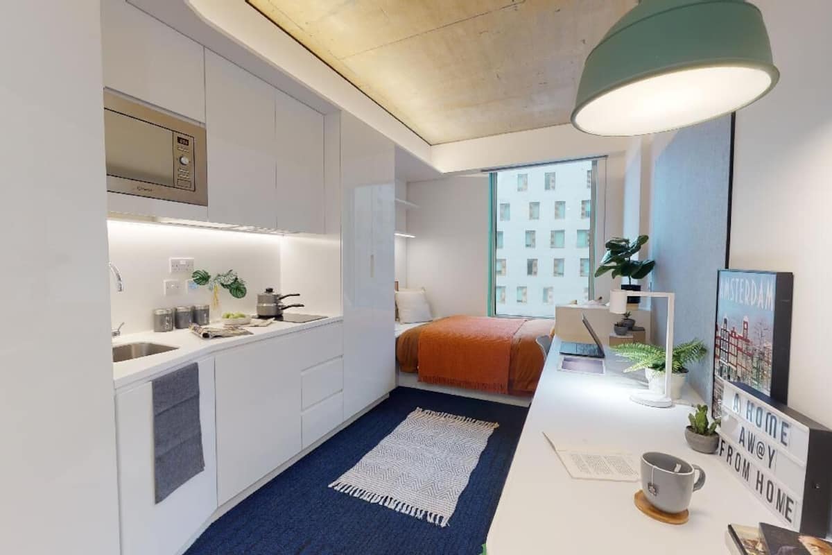 A compact hotel room with a kitchenette, a bed with an orange blanket, a blue rug, a desk with a lamp, and a cityscape poster.