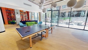Ein Pingpong-Tisch in einem modernen Innenraum mit großen Fenstern, einem grünen Sofa und einem Bücherregal voller Bücher.