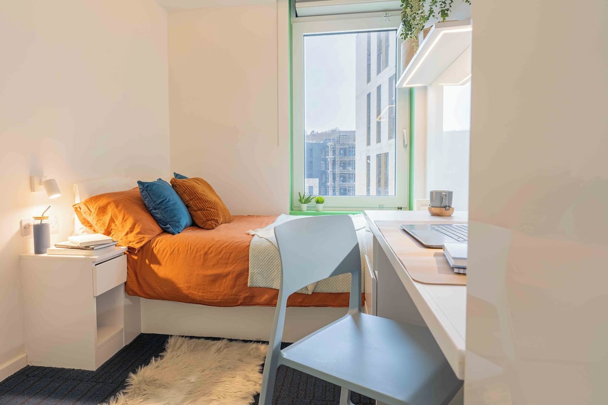 A bedroom with a bed, a desk, a chair, a nightstand, a lamp, a window, and a view of buildings.