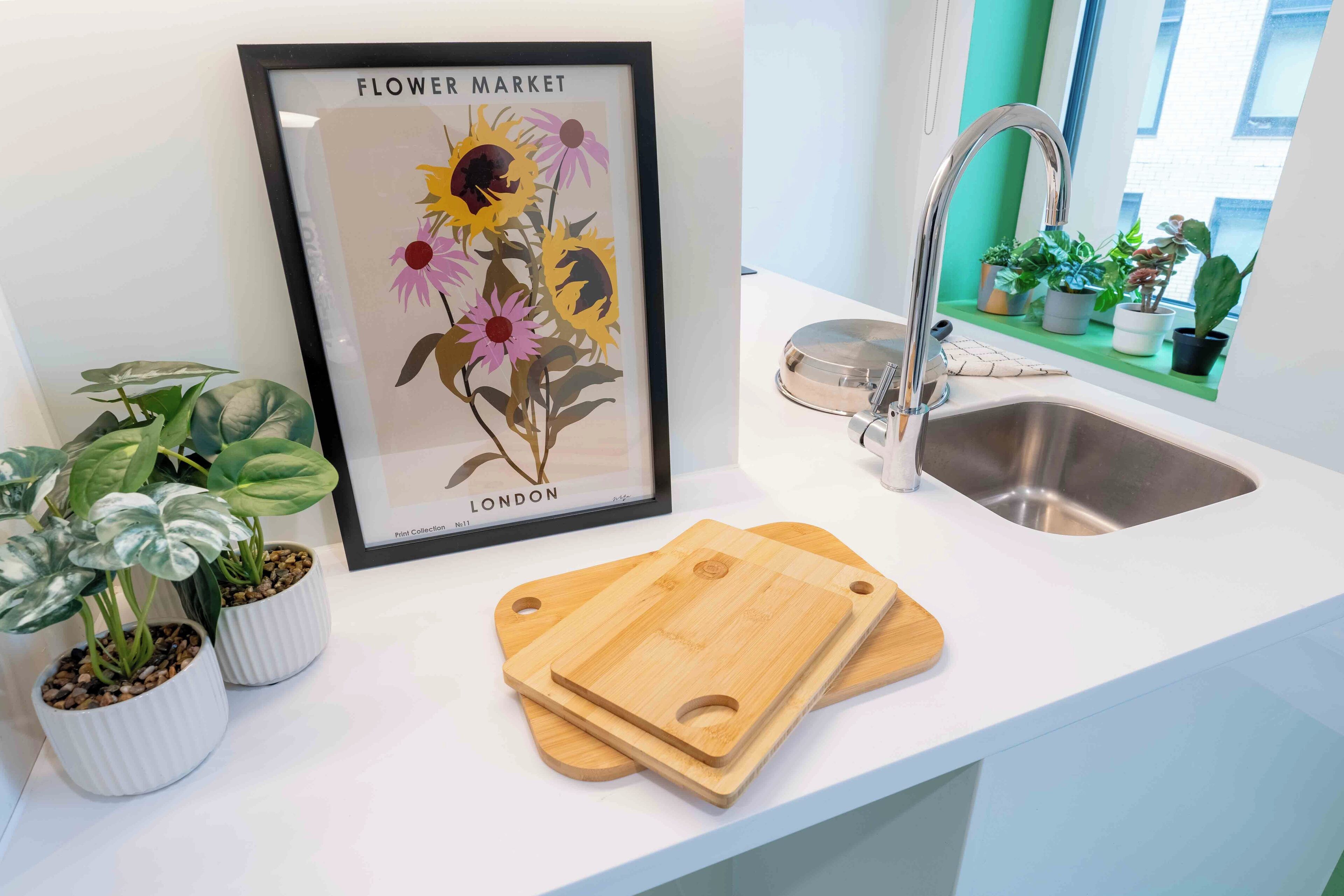 Een aanrecht met een poster van een bloemenmarkt, twee planten in potten, een spoelbak en houten snijplanken.
