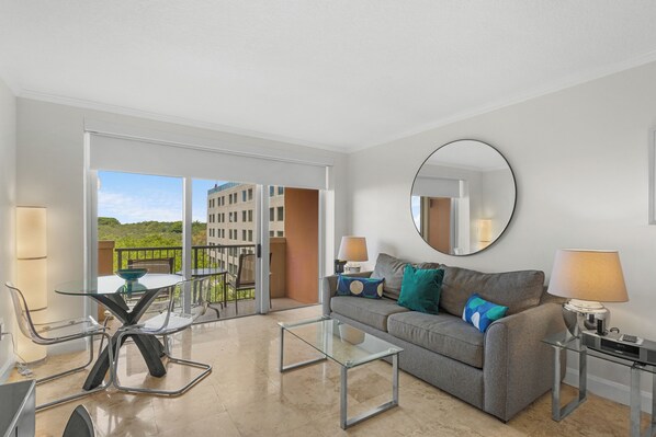 A modern living room with a glass coffee table, a grey sofa with cushions, a round mirror, and a balcony with a view of buildings and greenery.