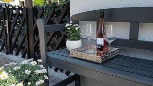 Un banc avec une bouteille de vin et des verres, une plante en pot aux fleurs blanches et un luminaire fixé au mur.