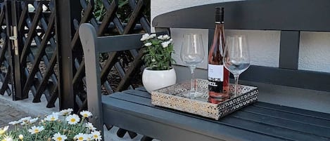 A bench with a bottle of wine and glasses, a potted plant with white flowers, and a wall-mounted light fixture.